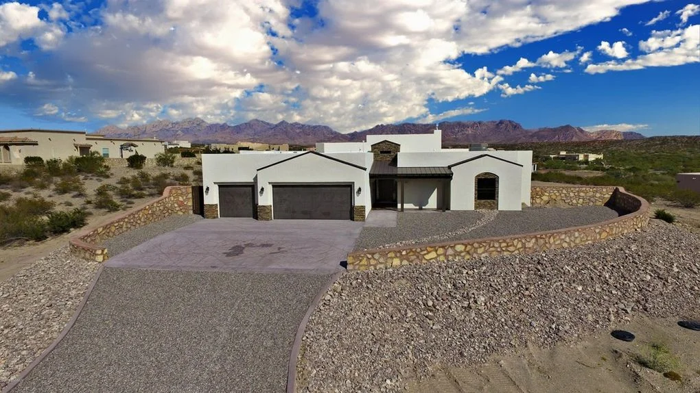 White stucco home with dark garage doors, stone accents, and gravel driveway in desert setting